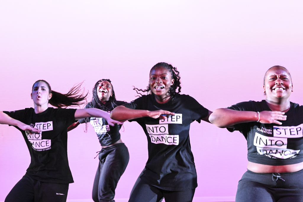4 dancers mid hip-hop movements all wearing black Step into Dance t-shirts and black bottoms. A wash of pink in the background.